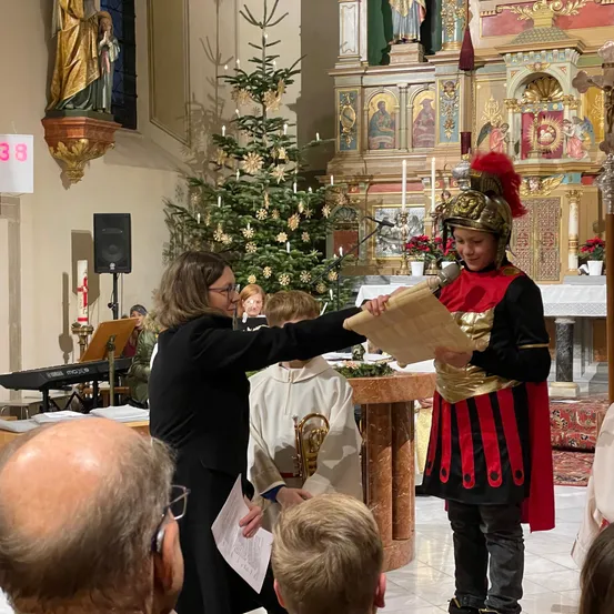 Eine Zeremonie in einer Kirche mit einem römischen Soldaten, der ein Schriftstück hält, und einer Person in einem schwarzen Gewand, die daraus liest, umgeben von Kindern und einem Publikum.