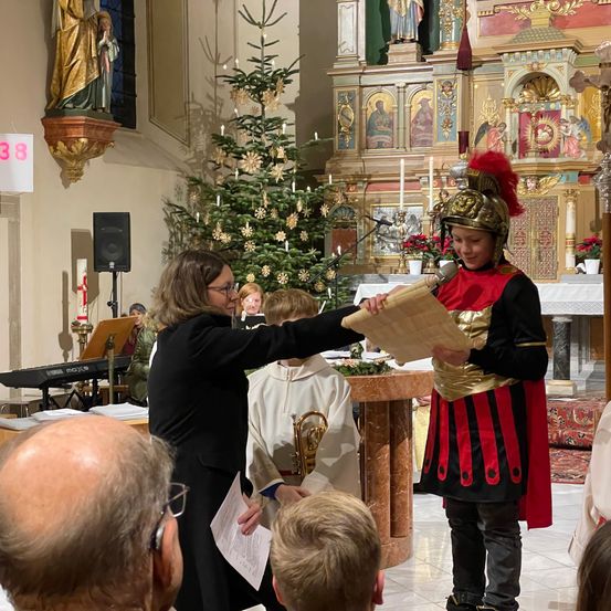 Eine Zeremonie in einer Kirche mit einem römischen Soldaten, der ein Schriftstück hält, und einer Person in einem schwarzen Gewand, die daraus liest, umgeben von Kindern und einem Publikum.