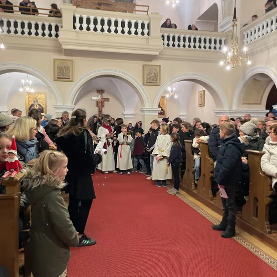 Eine Gemeinde versammelt sich in einer Kirche mit einem roten Teppich, der zum Altar führt. Menschen in weißen Gewändern spielen Instrumente. Ein Erwachsener steht und liest, während andere in Kirchenbänken stehen und sitzen.
