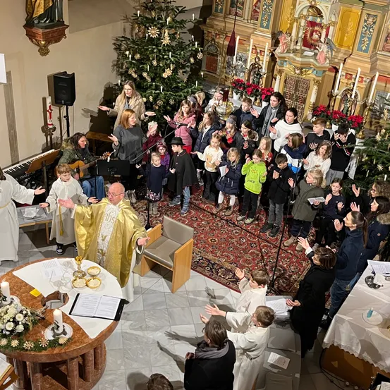 Eine Kirchenmesse mit einem Priester und Chor, umgeben von Erwachsenen und Kindern, vor einem geschmückten Weihnachtsbaum.