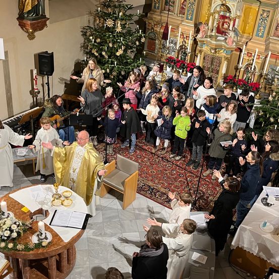 Eine Kirchenmesse mit einem Priester und Chor, umgeben von Erwachsenen und Kindern, vor einem geschmückten Weihnachtsbaum.