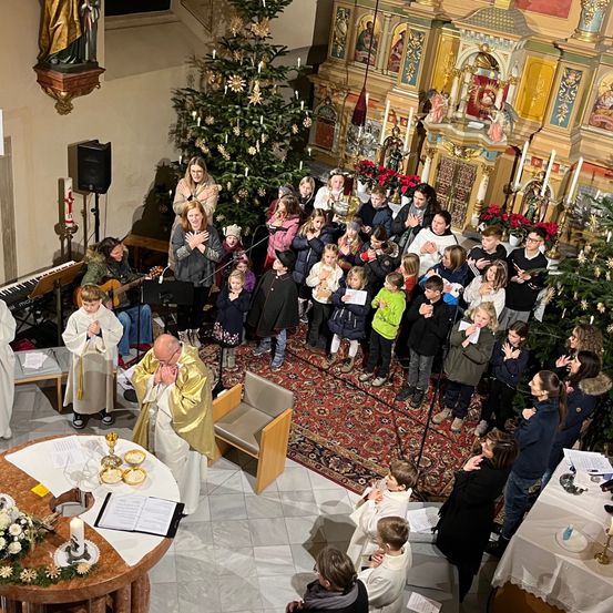 Ein Gottesdienst mit einem Priester und Chor, Kinder und Erwachsene beten vor einem Weihnachtsbaum. Es gibt einen geschmückten Altar mit Kerzen und Musikinstrumenten.