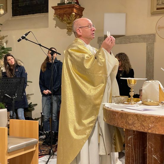 Ein Priester in einer goldenen Robe hält eine Hostie in einer Kirche. Hinter ihm stehen Menschen, darunter eine Frau mit einem Mikrofon. Der Altar hat einen Kelch, Schüsseln und ein Tuch.