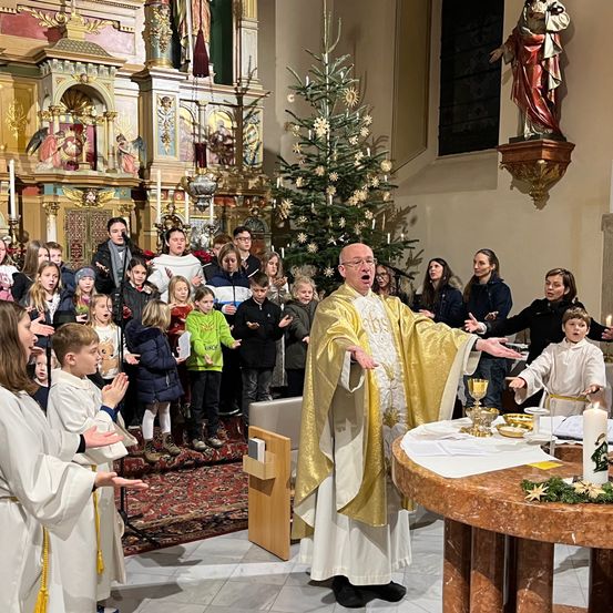 Ein Priester in einem goldenen Gewand leitet einen Kinderchor in einer Kirche an, mit einem Weihnachtsbaum im Hintergrund.