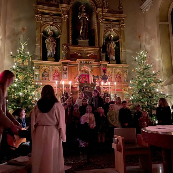 Ein Chor von Kindern in weißen Gewändern singt vor einem Altar, umgeben von zwei Weihnachtsbäumen und brennenden Kerzen. Eine Person in einem Pelzmantel spielt links die Gitarre.
