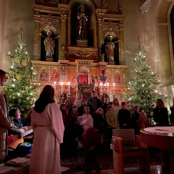 Eine Gruppe von Kindern und Erwachsenen singt Weihnachtslieder in einer Kirche, die mit Weihnachtsbäumen und Kerzen geschmückt ist. Eine Frau in einem weißen Gewand steht vor dem Chor.