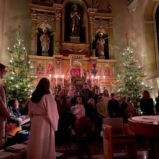 Eine Gruppe von Kindern und Erwachsenen singt Weihnachtslieder in einer Kirche, die mit Weihnachtsbäumen und Kerzen geschmückt ist. Eine Frau in einem weißen Gewand steht vor dem Chor.