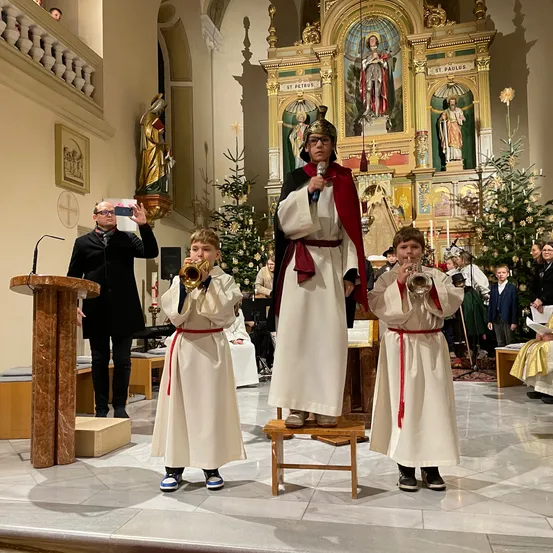 Zwei Jungen in weißen Gewändern spielen Trompeten in einer Kirche. Ein Mann in einem Kostüm steht zwischen ihnen. Ein Mann in einem schwarzen Anzug macht ein Foto.