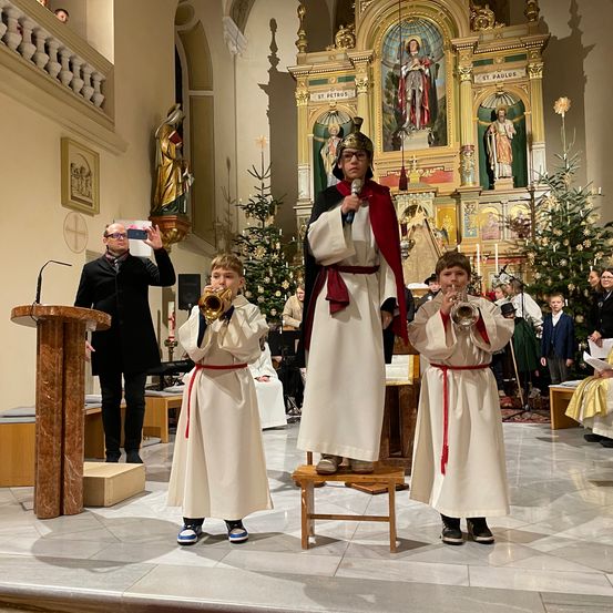 Zwei Jungen in weißen Gewändern spielen Trompeten in einer Kirche. Ein Mann in einem Kostüm steht zwischen ihnen. Ein Mann in einem schwarzen Anzug macht ein Foto.