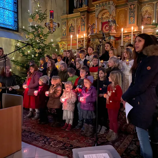 Eine Gruppe von Kindern mit Kerzen steht in einer Kirche. Eine Frau steht vorne und hält ein Mikrofon. Weihnachtsdekoration ist im Hintergrund zu sehen.