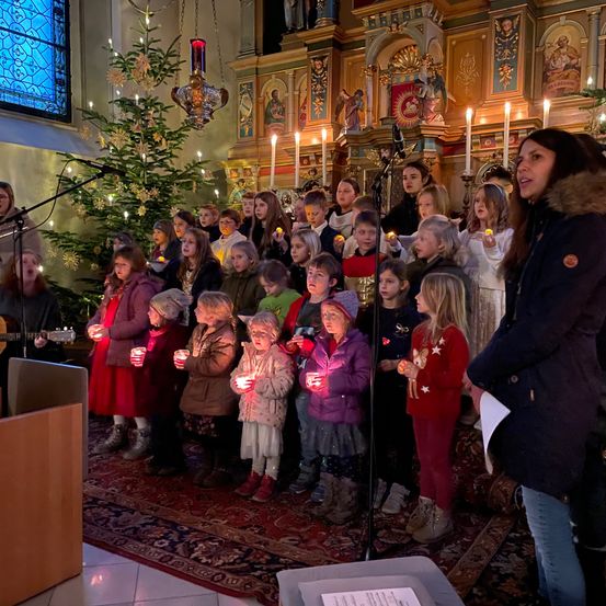 Eine Gruppe von Kindern mit Kerzen steht in einer Kirche. Eine Frau steht vorne und hält ein Mikrofon. Weihnachtsdekoration ist im Hintergrund zu sehen.