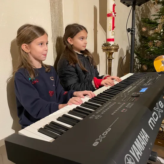 Zwei junge Mädchen spielen ein Yamaha-Piano in einem Raum mit einem Weihnachtsbaum und einer Kerze auf einem Kerzenständer im Hintergrund.