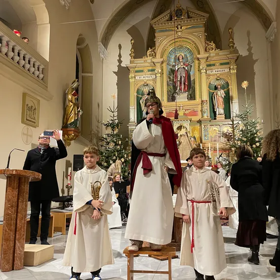 Ein Mann mit Helm steht auf einem Schemel in einer Kirche, begleitet von zwei Jungen mit Trompeten, während ein Mann dahinter ein Foto macht.