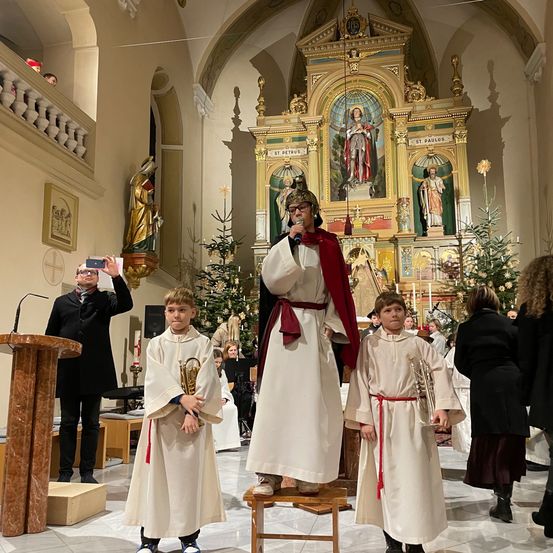 Ein Mann mit Helm steht auf einem Schemel in einer Kirche, begleitet von zwei Jungen mit Trompeten, während ein Mann dahinter ein Foto macht.