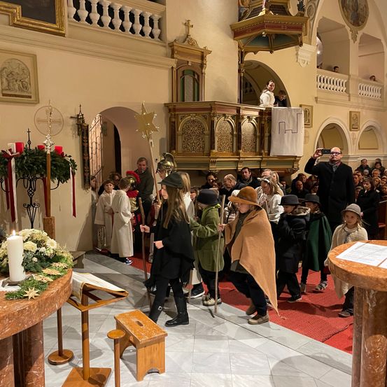 Eine Gruppe von Kindern in verschiedenen Kostümen geht in einer Kirche mit einem Altar, Kerzen und Blumen im Vordergrund.