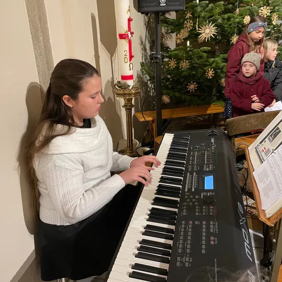 Ein Mädchen spielt Klavier vor einem Weihnachtsbaum mit einer Kerze und anderen Menschen um sie herum.