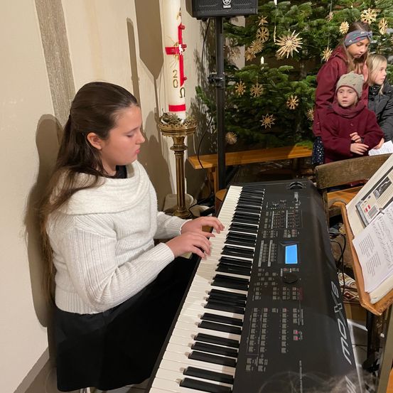 Ein Mädchen spielt Klavier vor einem Weihnachtsbaum mit einer Kerze und anderen Menschen um sie herum.