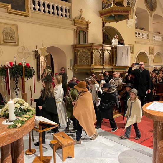 In einer Kirche gehen eine Gruppe von Menschen in traditioneller Kleidung an einem Altar vorbei, während ein Mann ein Foto macht und eine Menge von der Galerie zuschaut.