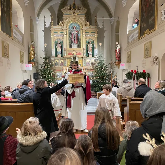 In einer Kirche spricht ein Mann in zeremonieller Kleidung in ein Mikrofon, während ein Junge eine Trompete hält. Die Menschen sitzen und stehen um ihn herum und beobachten.