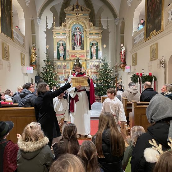 In einer Kirche spricht ein Mann in zeremonieller Kleidung in ein Mikrofon, während ein Junge eine Trompete hält. Die Menschen sitzen und stehen um ihn herum und beobachten.