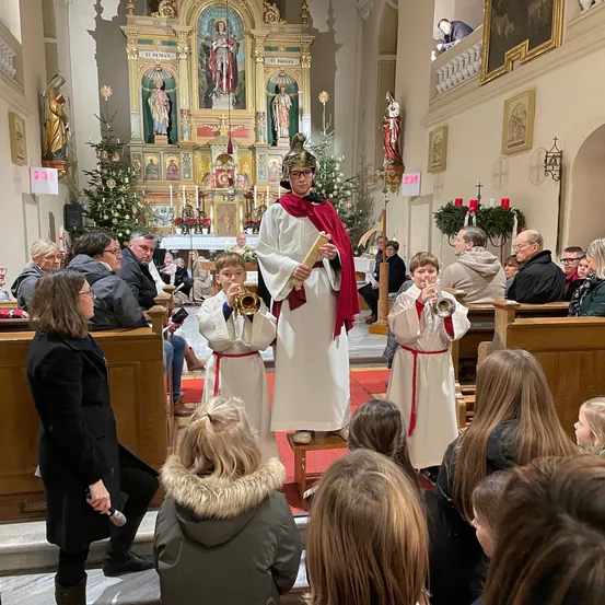 In einer Kirche spielen ein Priester und zwei Kinder Trompeten vor einem Publikum. Der Priester trägt einen roten Umhang und eine Brille. Die Kinder sind in weißen Gewändern.