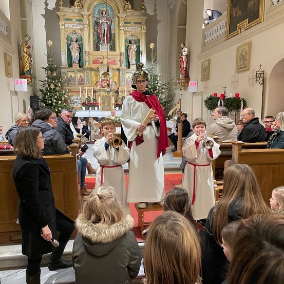 In einer Kirche spielen ein Priester und zwei Kinder Trompeten vor einem Publikum. Der Priester trägt einen roten Umhang und eine Brille. Die Kinder sind in weißen Gewändern.