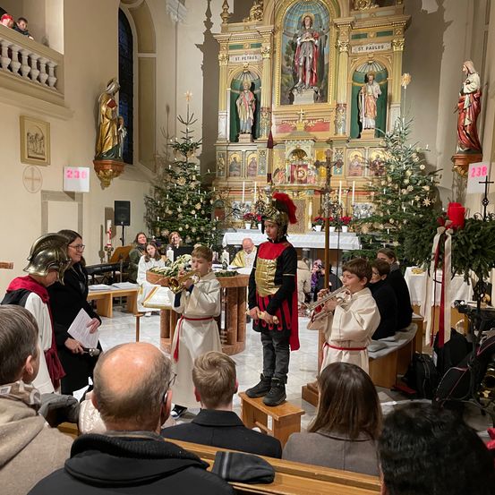 Eine Gruppe von Kindern in historischen Kostümen spielt Trompeten in einer Kirche. Sie stehen vor einem Altar mit Statuen und Weihnachtsbäumen. Dahinter sitzen Erwachsene in Kirchenbänken und schauen zu.