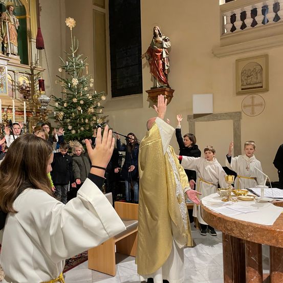 Ein Priester in einer Kirche leitet einen Kinderchor in einem festlichen Weihnachtsgottesdienst. Ein Weihnachtsbaum ist hinter dem Altar geschmückt.