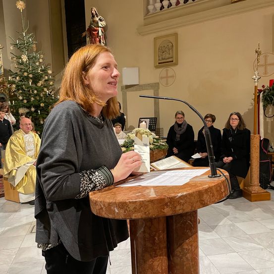 Eine Frau steht an einem Podest in einer Kirche und spricht eine sitzende Zuhörerschaft an. Hinter ihr sind ein Priester in einem gelben Gewand und ein Weihnachtsbaum zu sehen.