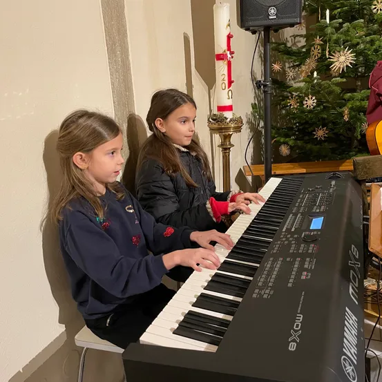 Zwei junge Mädchen sitzen an einem Klavier in einem Raum mit einem Weihnachtsbaum und einer brennenden Kerze.