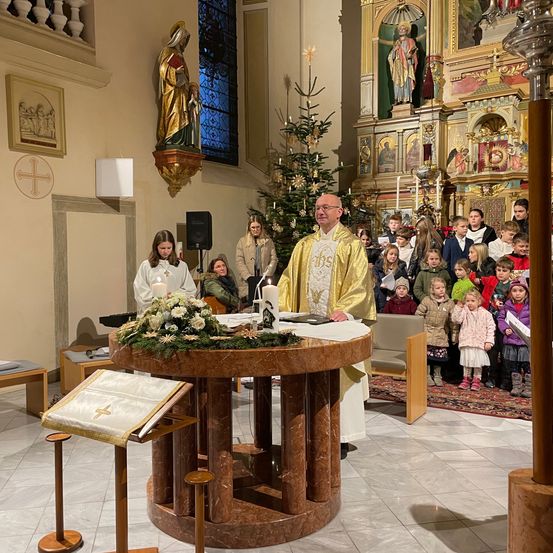 Ein Priester steht vor einem geschmückten Altar in einer Kirche mit Kindern und Erwachsenen. Kerzen, Blumen und ein Weihnachtsbaum sind vorhanden.