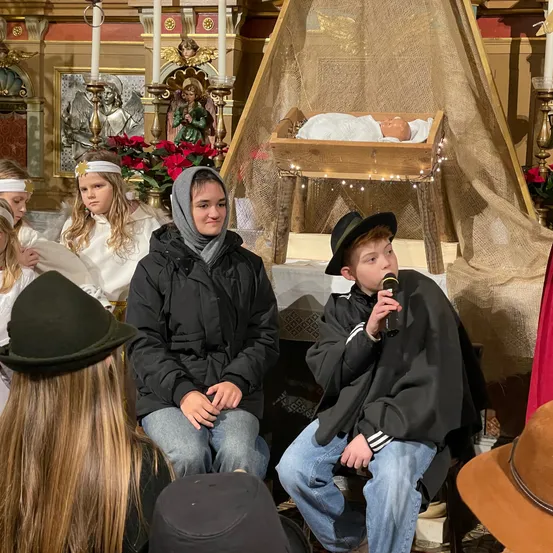 Mehrere Kinder sitzen in einem Raum mit einer Krippe im Hintergrund. Ein Junge hält ein Mikrofon, während ein Mädchen in einer Kapuze neben ihm sitzt.