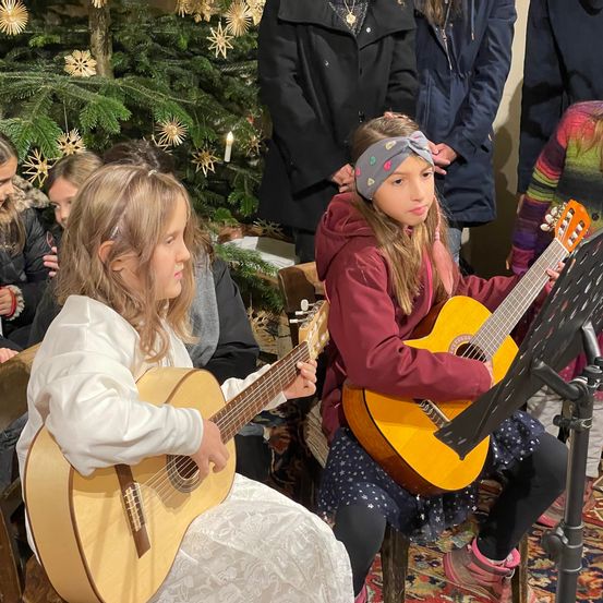 Mehrere junge Mädchen spielen Gitarren in einem für Weihnachten geschmückten Raum. Ein Mädchen trägt ein weißes Kleid, während ein anderes eine rote Kapuzenjacke und eine Stirnband trägt. Hinter ihnen sehen Erwachsene zu, und ein Weihnachtsbaum ist mit Ornamenten geschmückt.