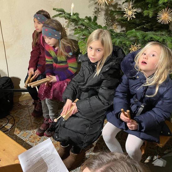 Vier junge Mädchen sitzen vor einem geschmückten Weihnachtsbaum. Jedes von ihnen hält einen Holzstab und scheint zu singen oder an einer festlichen Aktivität teilzunehmen.