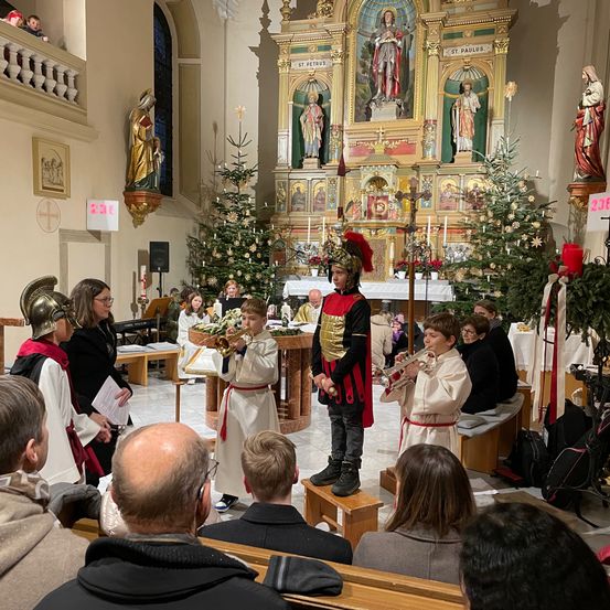 Eine Gruppe von Kindern in Kostümen spielt Trompeten vor einem Altar, während mehrere Menschen auf Kirchenbänken zusehen.