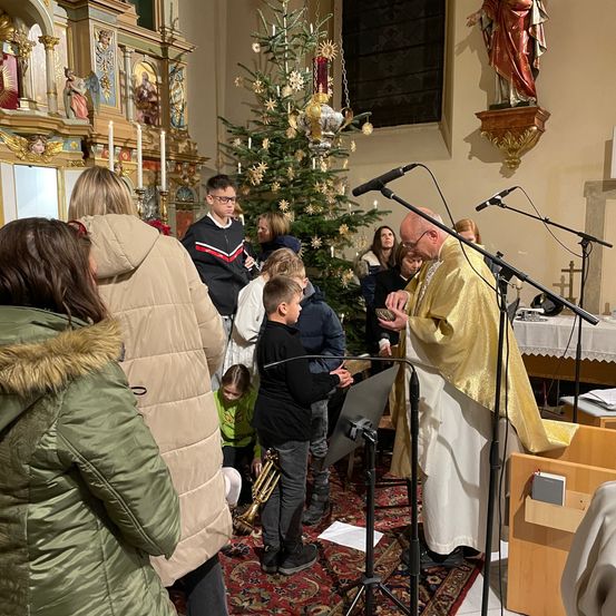 Ein Priester in einer Kirche liest aus einem Buch, umgeben von Mikrofonen und einem Weihnachtsbaum. Ein Junge hält eine Trompete und ein anderer ein Buch. Ein Mädchen kniet und eine Frau schaut den Priester an.