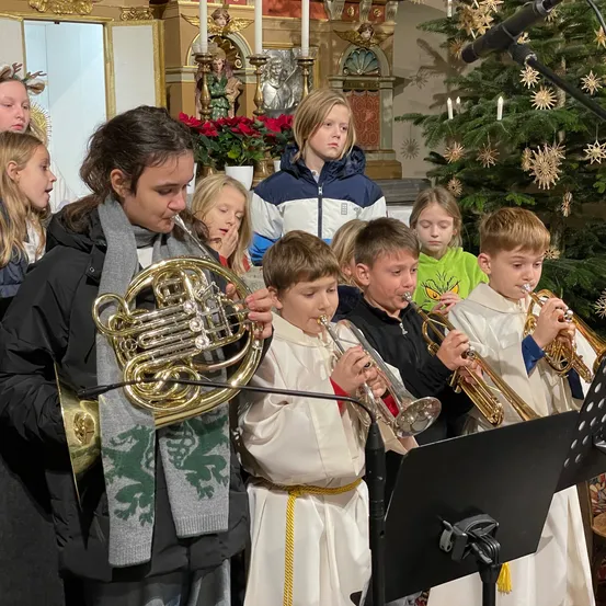 Eine Gruppe von Kindern in weißen Gewändern spielt Musikinstrumente in einer Kirche. Sie stehen in der Nähe eines Weihnachtsbaums und eines Mikrofons.
