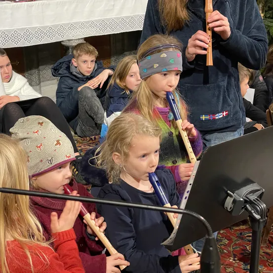Eine Gruppe von Kindern sitzt auf dem Boden und spielt Holzflöten vor einem Notenständer. Ein Mädchen trägt eine Stirnband und eine andere eine Mütze. Eine Frau in einer blauen Jacke hält eine Flöte. Hinter ihnen sitzt ein Junge auf einer Couch.