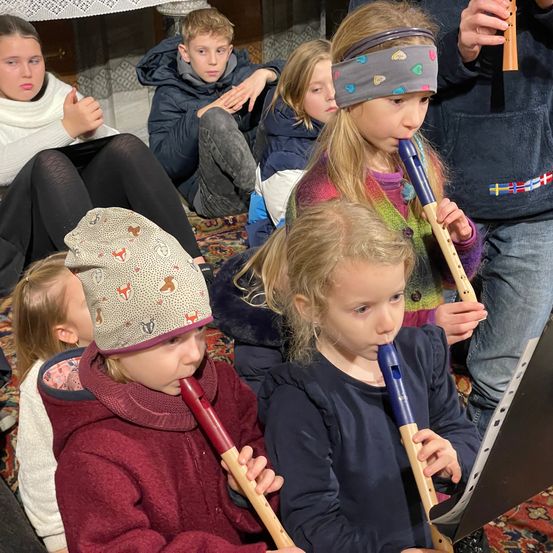 Mehrere Kinder spielen Blockflöten, während sie auf einem Teppichboden sitzen. Ein Mädchen mit Stirnband liest Musiknoten.