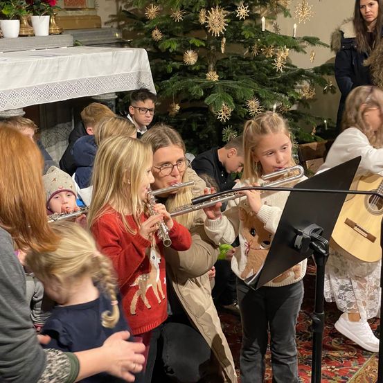 Eine Gruppe von Kindern und Erwachsenen versammelt sich in einem Raum mit einem geschmückten Weihnachtsbaum. Die Kinder spielen Flöten und werden von einer Frau begleitet, die eine Gitarre spielt.