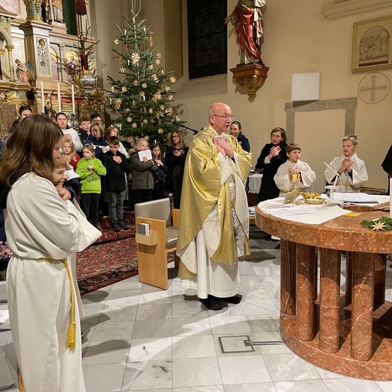 Ein Priester leitet eine religiöse Zeremonie in einer Kirche, umgeben von Kindern und Erwachsenen, mit einem geschmückten Altar und einem Weihnachtsbaum im Hintergrund.
