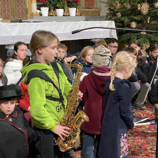 Mehrere Kinder sind um einen Jungen versammelt, der in einer Kirche Saxophon spielt. Einige sitzen, andere stehen, und die Umgebung ist festlich mit einem geschmückten Weihnachtsbaum im Hintergrund.