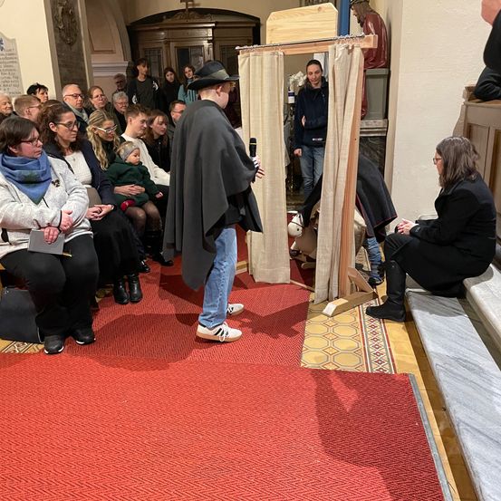 Eine Gruppe von Menschen ist in einer Kirche versammelt. Ein Mann steht vor einer Kabine mit Vorhängen und hält ein Mikrofon. Dahinter ist eine Statue zu sehen. Mehrere Menschen sitzen und stehen.