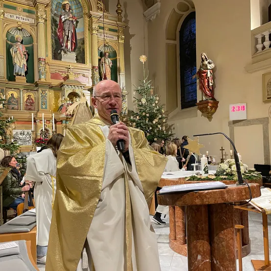 Ein Priester in einem goldenen Gewand steht an einem Podium und hält ein Mikrofon. Hinter ihm befindet sich ein Altar mit einem geschmückten Weihnachtsbaum. In der Nähe sitzen und stehen Menschen.