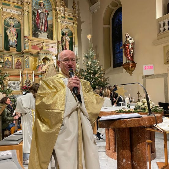 Ein Priester in einem goldenen Gewand steht an einem Podium und hält ein Mikrofon. Hinter ihm befindet sich ein Altar mit einem geschmückten Weihnachtsbaum. In der Nähe sitzen und stehen Menschen.
