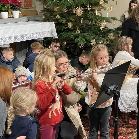 Kinder spielen Flöten in einer Kirche, umgeben von einem festlichen Weihnachtsbaum, und Erwachsene und Kinder beobachten sie aufmerksam.