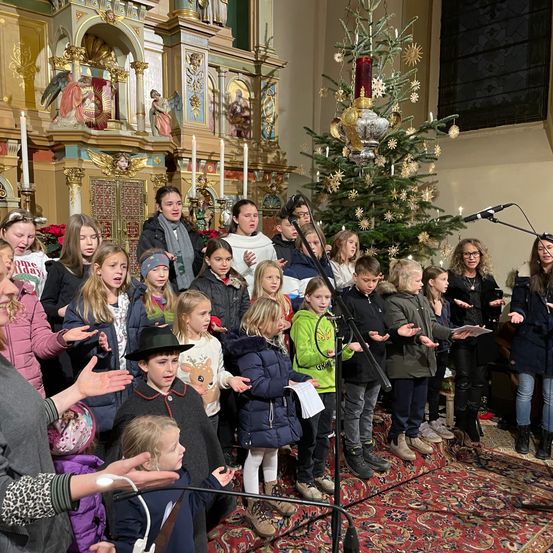 Ein Chor aus Kindern und Erwachsenen steht vor einem geschmückten Weihnachtsbaum und singt während eines Gottesdienstes in einer Kirche.
