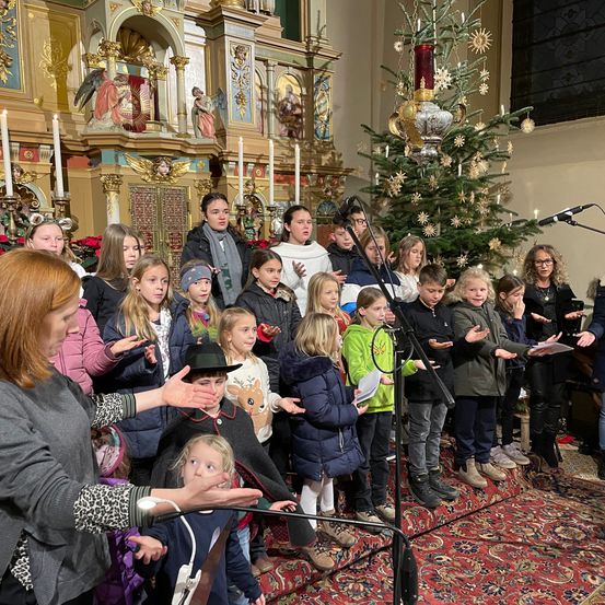 Ein Kinderchor in Winterkleidung singt vor einem geschmückten Weihnachtsbaum. Sie stehen auf einem Teppich mit einem Mikrofonständer, in der Nähe einer Frau, die ein Blatt Papier hält.