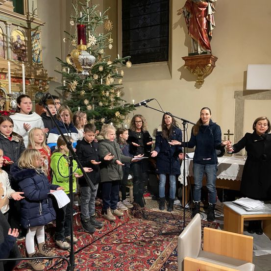 Eine Gruppe von Kindern und Erwachsenen versammelt sich in einer Kirche, um gemeinsam zu singen. Sie stehen vor einem geschmückten Weihnachtsbaum, mit Mikrofonen auf Ständern und einem Teppich auf dem Boden.