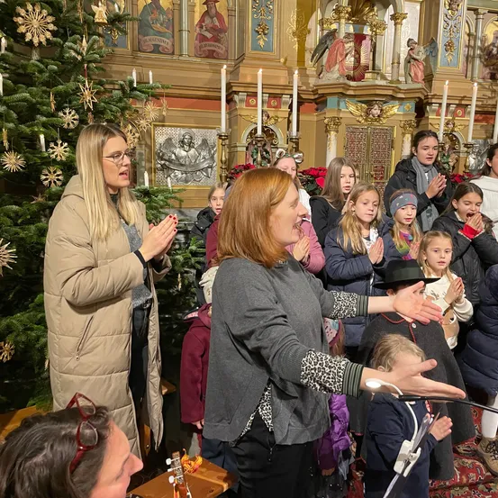 Ein Kinderchor singt in einer für Weihnachten geschmückten Kirche. Eine Frau mit Brille leitet sie.